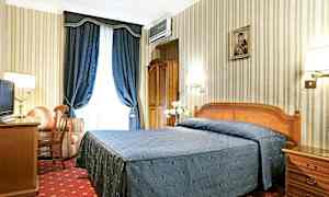 Et hotellrom med en dobbeltseng dekket med et blått sengeteppe, et skrivebord, en lenestol og blå gardiner. (AI-generert tekst)