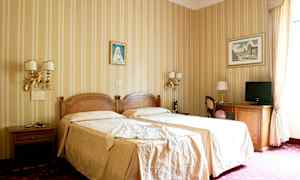 Et hotellrom med to enkeltsenger dekket med beige sengetøy, dekorerte vegger med malerier, og møbler inkludert bord, stoler og en TV. (AI-generert tekst)