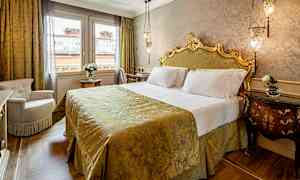 Et elegant soverom med en dekorativ seng med hodegjerde, dekket med et gyllent sengeteppe. Rommet har to vinduer og et bord med en blomsterpotte. (AI-generert tekst)