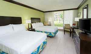 Et hotellrom med to dobbeltsenger, et skrivebord med en TV, og store glassdører som vender ut mot en hage. (AI-generert tekst)