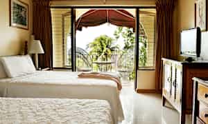 Et hotellrom med to enkeltsenger, en TV på en trekommode og en åpning til en balkong med utsikt over vegetasjon og palmetrær. (AI-generert tekst)