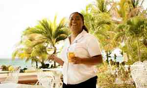 En person holder to glass, ett med vin og ett med juice, ved et bord utendørs nær stranden og omgitt av palmer. (AI-generert tekst)