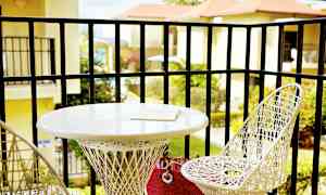 En balkong med to hvite flettede stoler og et rundt bord. På bordet ligger en bok. Utsikt over beplantning og bygninger i bakgrunnen. (AI-generert tekst)