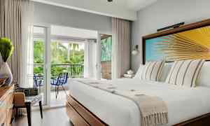 Hotellrom med en hvit seng, dekorativ veggkunst med palmeblad, utsikt mot en balkong med frodig natur. (AI-generert tekst)