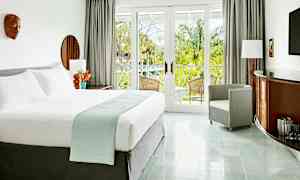 Et lyst og elegant hotellrom med en stor seng, lenestol og møbler med tredetaljer. Rommet har store glassdører som leder til en balkong med utsikt over frodige palmer. (AI-generert tekst)