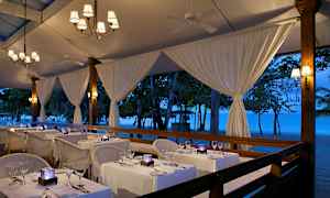 En restaurant med bord dekket med hvite duker ved stranden under et kveldspreget telt. (AI-generert tekst)