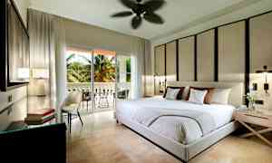 Et moderne soverom med stor seng, dekorativ veggpanel og åpning til en balkong med palmer utenfor. (AI-generert tekst)