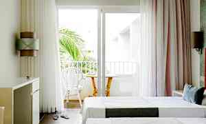 Lyst hotellrom med to enkeltsenger og en balkong med stoler og bord. (AI-generert tekst)