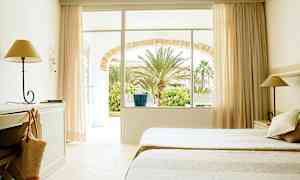 Et hotellrom med to senger, skrivebord og lampe. Vinduer og dører leder ut til en hage med palmer. (AI-generert tekst)