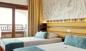 Et hotellrom som viser to enkeltsenger med turkise dekorasjoner, bak sengene er det en vegg med kunstnerisk dekorasjon og en liten lampe. (AI-generert tekst)