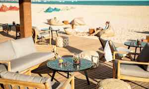 Terrasse med sittemøbler og bord, i bakgrunnen en sandstrand og havet. (AI-generert tekst)