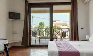 Et rom med en seng, en liten TV på veggen og en balkong med utsikt over bygninger og palmer. (AI-generert tekst)