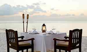 Et oppdekket bord er plassert på en sandstrand nær vannet med lykter og fakler ved siden av. (AI-generert tekst)