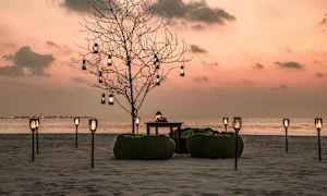 En strandmiljø ved solnedgang med et dekorert tre, sittesekker og bord samt lange lykter. (AI-generert tekst)