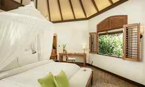 Et lyst rom med et hvitt tak, en seng med myggnett og grønne puter, et skrivebord og en stol ved et vindu med utsikt over vegetasjon. (AI-generert tekst)