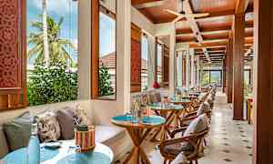 Bilde av en restaurant med bord og stoler, dekorert med puter og utsikt mot vegetasjon gjennom vinduene. (AI-generert tekst)