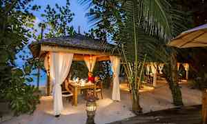 En opplyst uteplass med tak og hvite gardiner, omgitt av grøntområde på en strand. (AI-generert tekst)