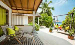 Et uteområde med trestoler, en huske og sand, omgitt av grøntområder og palmetrær. (AI-generert tekst)