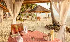 Bord dekket med servietter og tallerkener på sanden ved kysten, omgitt av tak med tekstildekorasjoner. (AI-generert tekst)