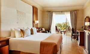 Et hotellrom med stor seng, skrivebord og balkong med utsikt over grøntområde. (AI-generert tekst)