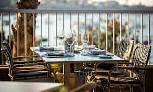 Et dekket bord med glass og servietter på en utendørs terrasse, omgitt av utsikt over vannet. (AI-generert tekst)