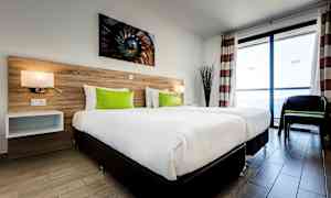 Hotellrom med to enkeltsenger, hvitt sengetøy og grønne puter, en sengegavl av trepanel og store vinduer som slipper inn naturlig lys. (AI-generert tekst)