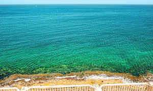 En utsikt over et klart blått hav med en steinete strand, sett fra et sted med hvite rekkverk. (AI-generert tekst)