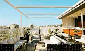 Bilde av en takterrasse med sittegrupper, bord og parasoller. Utsikt over bygninger og en klarblå himmel. (AI-generert tekst)