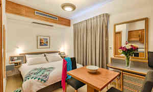 Et lyst hotellrom med en dobbeltseng, spisebord, sitteplasser og et speil. Dekorert med en blomsterpotte på bordet. (AI-generert tekst)