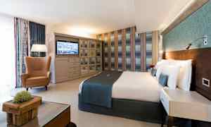 Et hotellrom med en dobbeltseng, en lenestol, et bord og en TV på veggen. (AI-generert tekst)