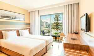 Et hotellrom med to enkeltsenger, et skrivebord og utsikt over en balkong med basseng og trær. (AI-generert tekst)