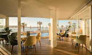 En restaurant med sitteplasser og glassvegger med utsikt over et basseng og palmer ved havet. (AI-generert tekst)