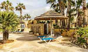 En steinlagt uteplass med solsenger og stråtekte parasoller omgitt av palmer og steinbygninger. (AI-generert tekst)