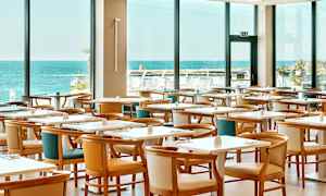 Restaurant med flere bord og stoler, stor vindusutsikt mot havet. (AI-generert tekst)