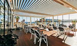 En utendørsterrasse med bord og stoler, omgitt av glassvegger og med utsikt over havet og palmer. (AI-generert tekst)