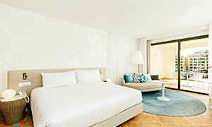 Et moderne hotellrom med en stor seng, lyse vegger og elegant innredning. Rommet har et sitteområde med puter samt en romslig balkong med utsikt. (AI-generert tekst)