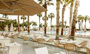 En utendørs restaurant med flere oppstilte bord og stoler, omgitt av palmer og med utsikt over et basseng. (AI-generert tekst)