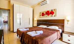 Et soverom med to enkeltsenger, brune sengetepper og hvitt sengetøy. På veggen over sengene henger et bilde med røde blomster. Til venstre finnes et garderobeskap med blank hvit overflate. (AI-generert tekst)