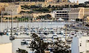 En marina med mange båter, omgitt av historiske bygninger og frodig vegetasjon. (AI-generert tekst)