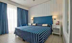 Et hotellrom med en dobbeltseng med blå- og hvitstripete sengetøy, en blå sengegavl og hvite møbler. (AI-generert tekst)