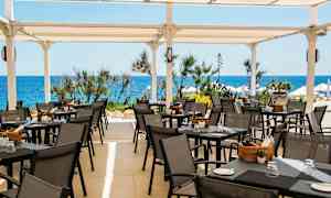 En terrasse med uteservering, bord og stoler under et hvitt tak, med havet og vegetasjon i bakgrunnen. (AI-generert tekst)