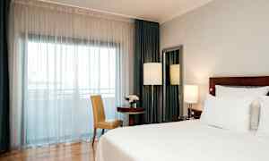 Et hotellrom med en stor seng, et bord med en stol, og et vindu dekket av hvite gardiner. (AI-generert tekst)