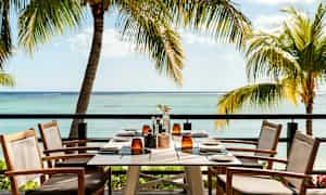 Et dekket bord med sitteplasser står på en terrasse med utsikt over havet og omkringliggende palmer. (AI-generert tekst)