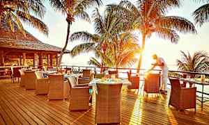 Uteservering med utsikt over havet og solnedgangen, palmetrær i bakgrunnen og en servitør som forbereder et bord. (AI-generert tekst)