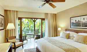 Et smakfullt innredet hotellrom med en stor seng, sittegruppe og en balkong med utsikt over frodige palmer. (AI-generert tekst)