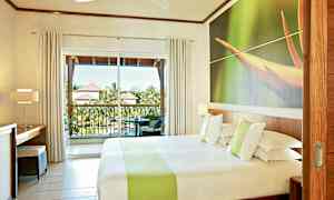 Hotellrom med en dobbeltseng, en stor vindusvegg og en balkong med utsikt over grønne områder og takene på nærliggende bygninger. (AI-generert tekst)