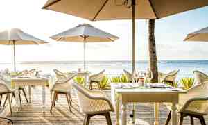 En utendørs restaurant med bord og parasoller ved sjøen. (AI-generert tekst)