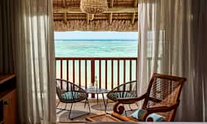 Utsikt over havet fra et rom med en overbygd balkong. Mellom gjennomsiktige gardiner er det to hagestoler ved et bord. (AI-generert tekst)