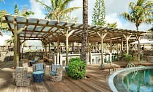 En lounge ved et basseng på et tropisk feriested, med sitteplaser og en bar under trebjelker. (AI-generert tekst)