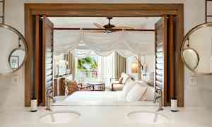 Et hotellrom med sittegruppe, seng og åpning mot en veranda. I forgrunnen ses et bad med dobbelt servant og speil. (AI-generert tekst)